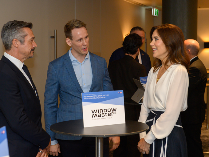 Bei einer Konferenz oder einer Networking-Veranstaltung stehen Menschen in Geschäftskleidung um einen Stehtisch mit der Aufschrift „WindowMaster“ herum und unterhalten sich.