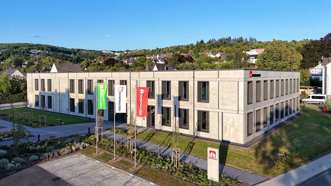 Modernes Betongebäude mit großen Fenstern, Fahnen und gepflegter Außenanlage vor dem Hintergrund grüner Hügel und blauem Himmel.
