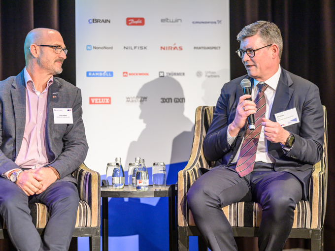 Zwei Männer in Anzügen unterhalten sich auf der Bühne, im Hintergrund sind ein Mikrofon, Wasserflaschen und eine Werbetafel zu sehen.