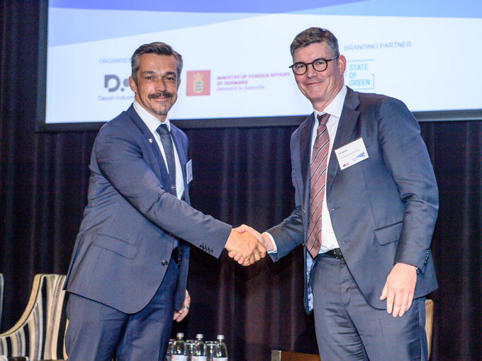 Zwei Männer in Anzügen, die sich bei einer offiziellen Veranstaltung die Hand geben, im Hintergrund eine Präsentationsleinwand und Wasserflaschen.