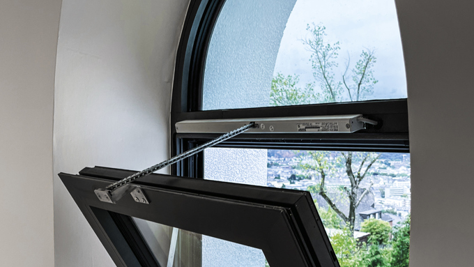 Ein modernes Bogenfenster steht einen Spalt breit offen und gibt den Blick auf Grünflächen und einen teilweise fertiggestellten Außenbereich dahinter frei.